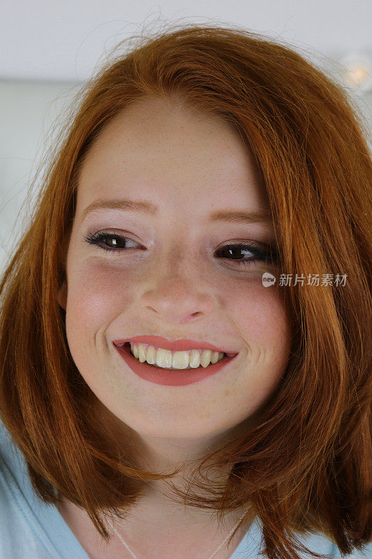 照片中，14 / 15岁的红发少女，皮肤苍白，脸上有雀斑，坐在卧室里看着镜头，神情轻松，面带微笑